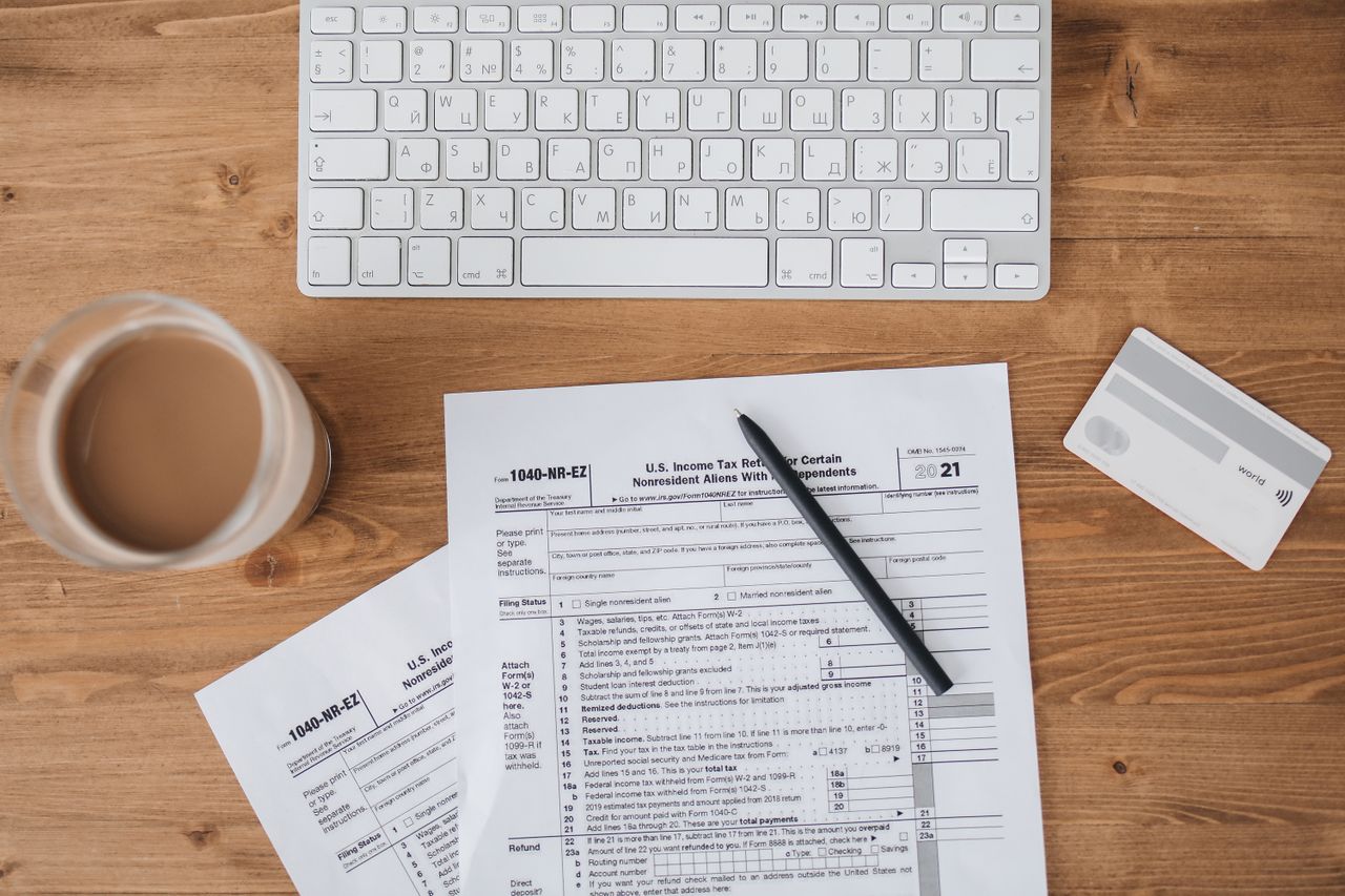 Top view of tax forms, keyboard, pen, and coffee on a wooden desk.